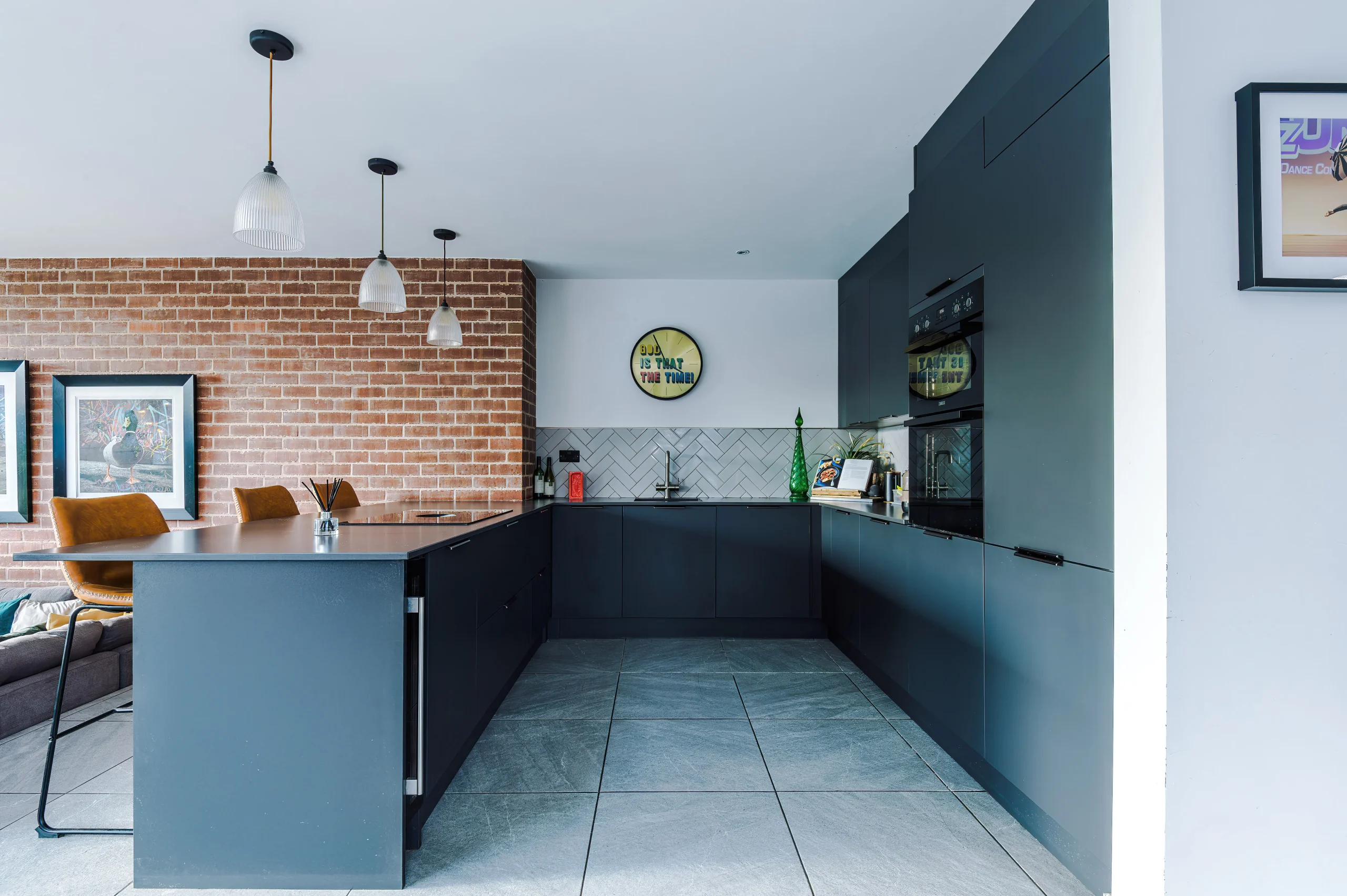 View of dark grey kitchen with exposed brick and breakfast bar overlooking lounge.