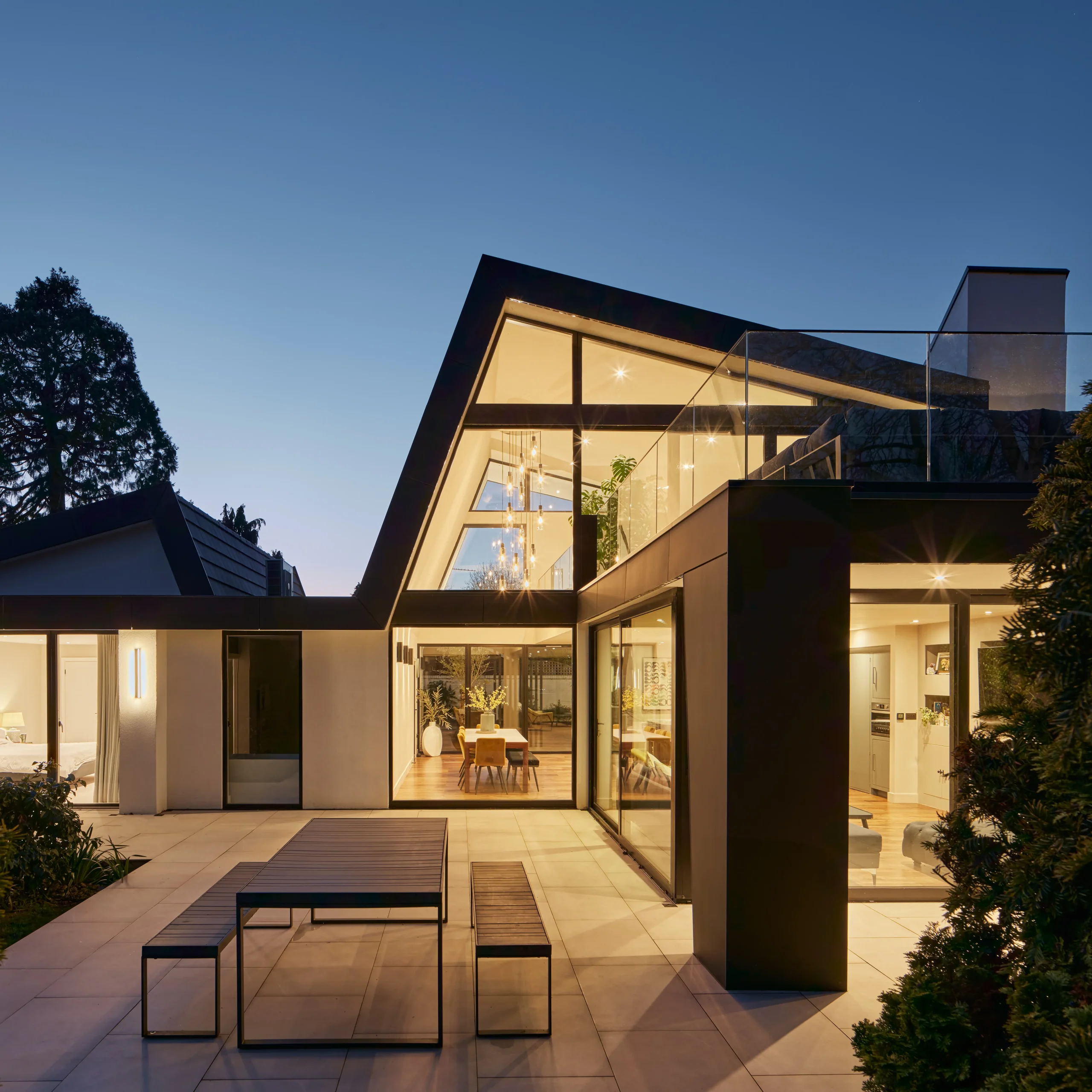 View of the double-height living and dining space with floor-to-ceiling glazing opening onto a paved terrace at the rear.