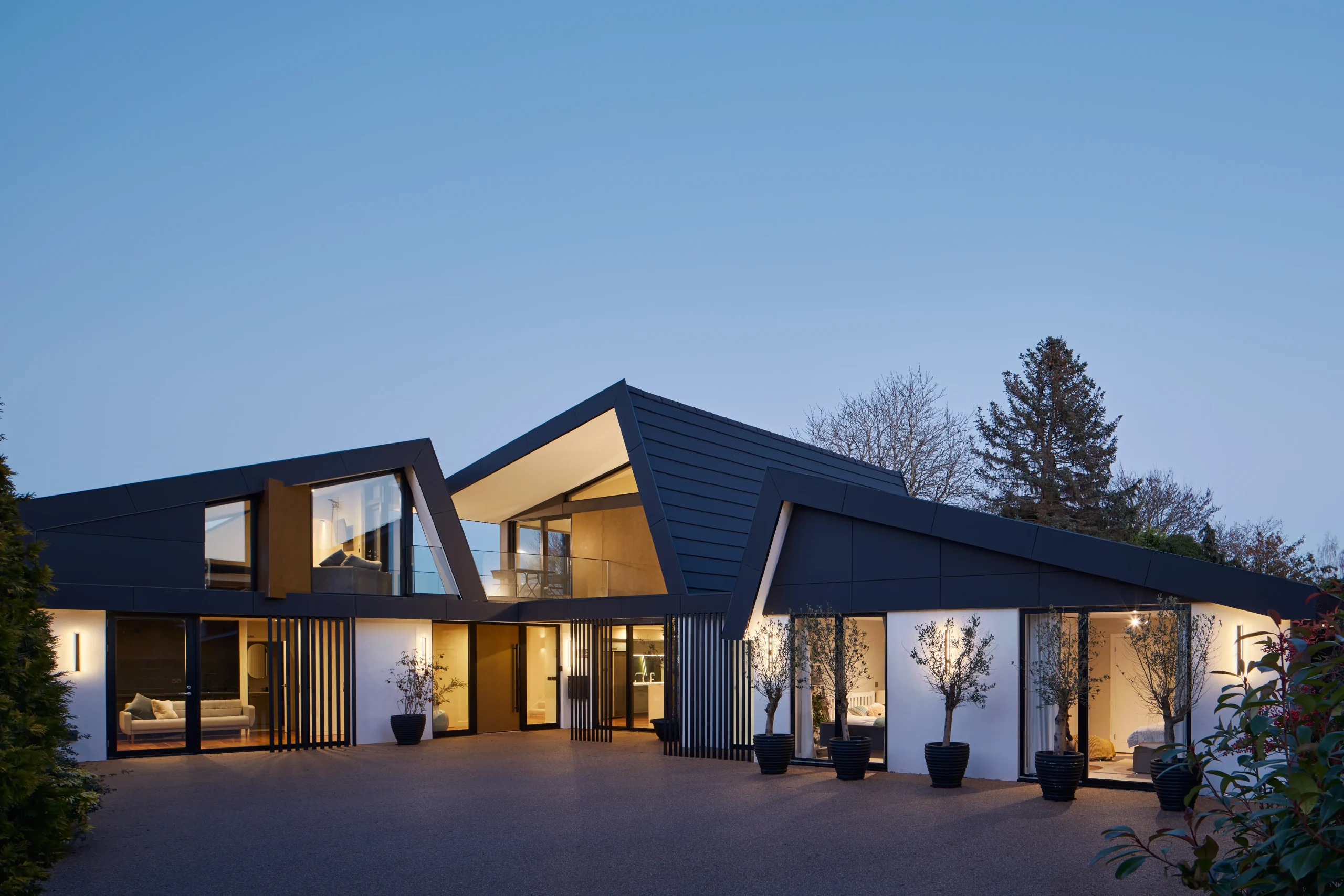 Wide dusk view showing single- and two-storey volumes arranged beneath mirrored roof forms with illuminated interiors