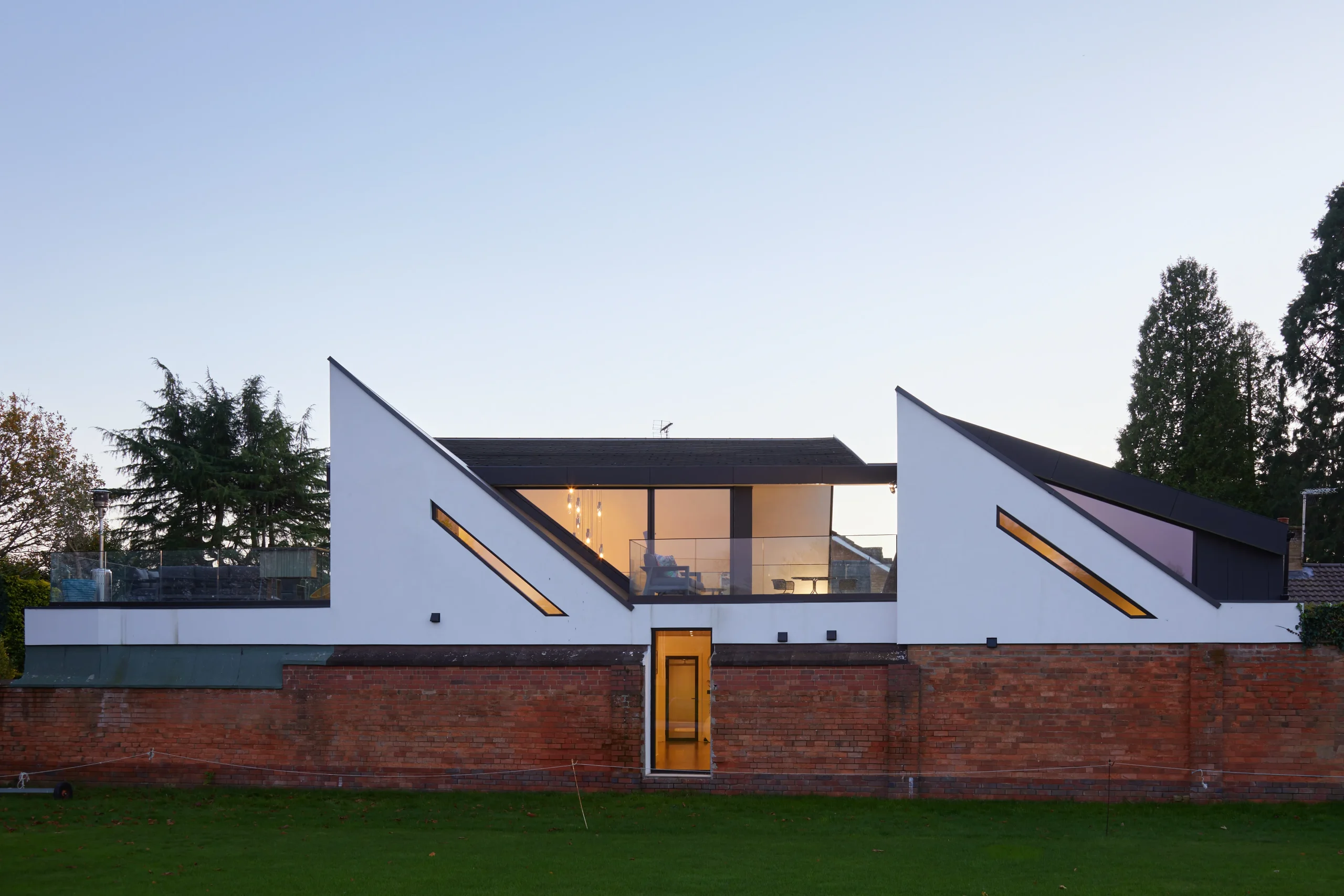 Viewed from the adjacent cricket ground, triangulated stair volumes and elevated decks rise above the brick boundary wall, framing views toward the pitch